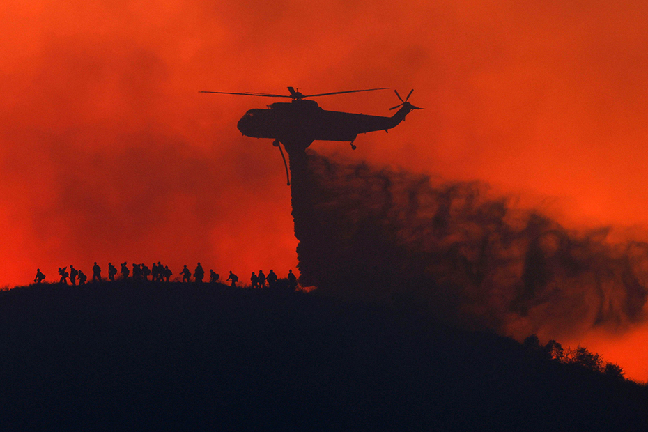 A water tanker drops water while firefighters walk below, as the Sandy Fire approaches in Simi Valley